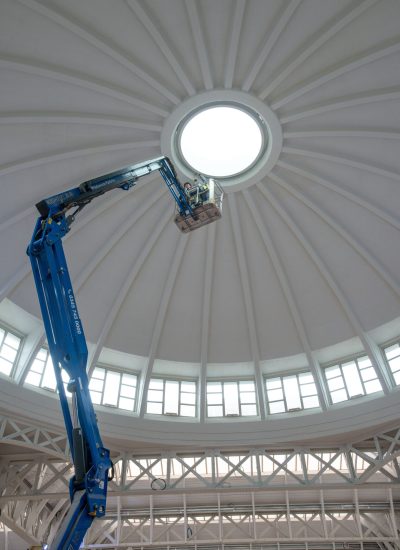 A worker in a blue lift performs maintenance near the central skylight of a large domed ceiling, with radial beams and windows around the base, high above the floor of this impressive London venue.
